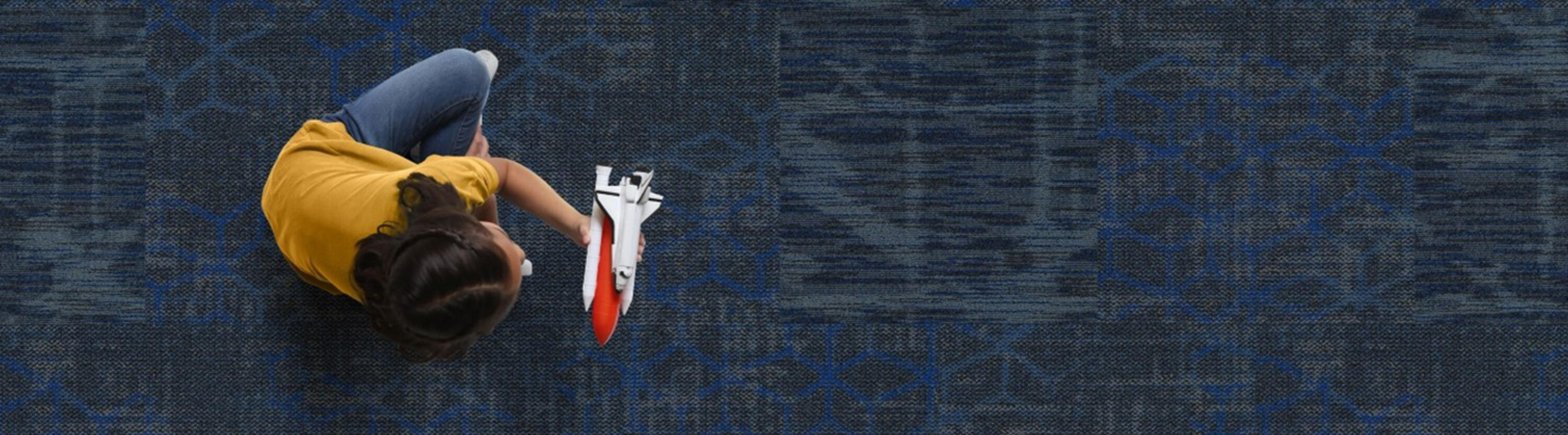 Young student sitting on blue carpeted floor in classroom, playing with rocket ship.